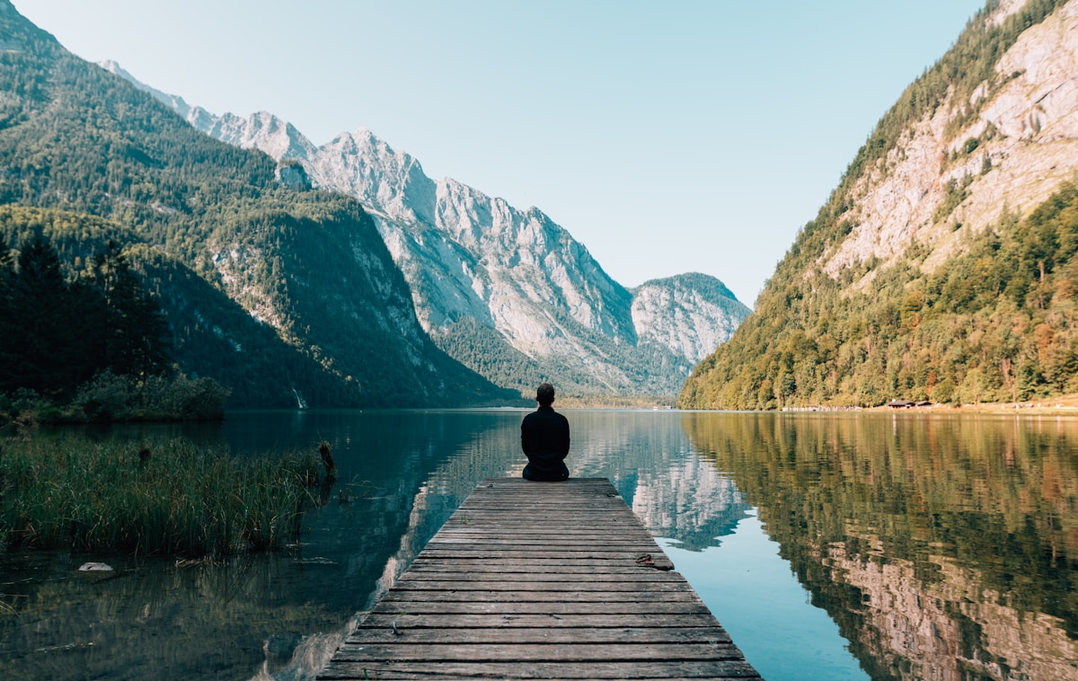 Person sitting alone on a dock facing still mountain lake — intergenerational trauma